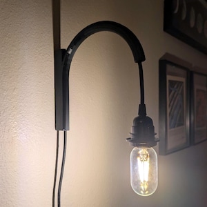 May include: Black wall-mounted light fixture with a curved arm and a clear glass bulb. The bulb is lit, emitting a warm glow. The fixture is mounted on a light-colored wall, with framed artwork visible in the background.