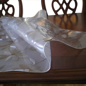 May include: A clear plastic table cover with a floral pattern. The cover is rolled up and placed on a wooden table.