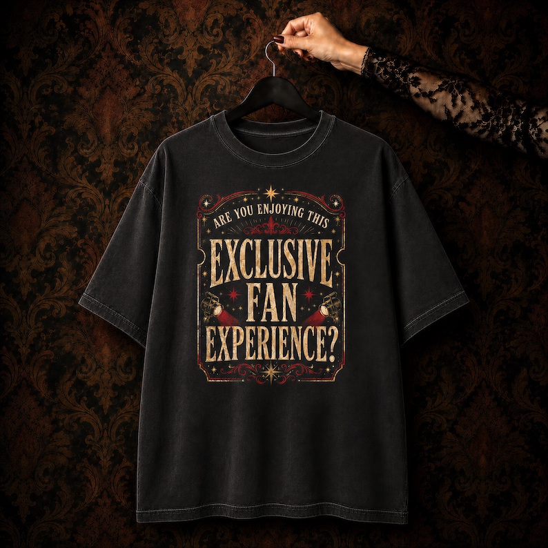 May include: A black short-sleeve t-shirt featuring a vintage-style graphic print in gold and red. The text reads: ARE YOU ENJOYING THIS EXCLUSIVE FAN EXPERIENCE? The design includes decorative flourishes, stars, and two small spotlight icons.