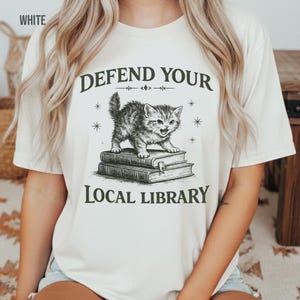 May include: Cream-coloured t-shirt with a graphic of a kitten on books, and the text "DEFEND YOUR LOCAL LIBRARY". The kitten is grey and white, and the text is in a dark, vintage font. The shirt is a casual style.