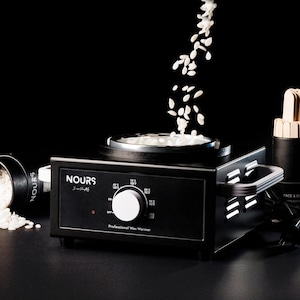 Puede incluir: Calentador de cera negro con un dial blanco y un recipiente transparente para la cera. El calentador tiene la palabra "NOURS" impresa en la parte delantera. Hay un recipiente de perlas de cera al lado del calentador y un recipiente de espátulas de madera a la derecha.