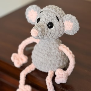 May include: A handmade crochet mouse toy in grey with pink accents. The mouse has large black eyes, a pink nose, and pink-lined ears. Its arms and legs are also pink. The toy is sitting on a dark brown surface.