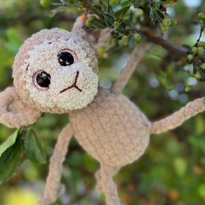 May include: A handcrafted monkey doll in beige and tan tones, suspended from a tree branch. The monkey features large, glittery eyes and a smiling expression. The soft, textured material gives it a cuddly appearance.