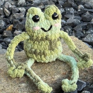 Könnte beinhalten: Eine handgefertigte Häkelfroschfigur in Grün- und Brauntönen, mit schwarzen Augen und einem lächelnden Mund. Der Frosch hat rosa Wangen und sitzt auf einem Stein. Das Plüschtier ist ein skurriles und charmantes Dekorationsobjekt.