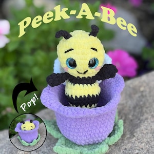 May include: A plush yellow and black bee toy emerging from a purple flower. The bee has large, sparkly blue eyes and black antennae. The text "Peek-A-Bee" is arched above the bee. A smaller inset image shows the bee inside the flower.