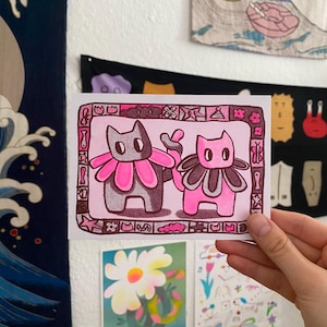May include: A rectangular art print featuring two stylized cats with large, petal-like collars. The left cat is dark gray with a bright pink collar, and the right cat is pink with a dark gray collar. The border contains small, varied icons.