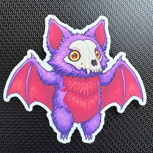 May include: A sticker featuring a cartoon bat with a skull-like face, purple fur, and red wings. The bat has yellow eyes and a fluffy red chest. The sticker is on a black textured surface.