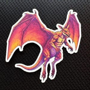 May include: A sticker featuring a stylized illustration of a winged kangaroo-like creature. The creature has a muscular build, with a red, orange, and purple color scheme. The wings are spread, and the creature appears to be in mid-flight.