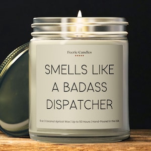 May include: A clear glass jar candle with a silver lid, containing white wax and a lit wick. The label displays the text Feerie Candles, five stars, and the phrase SMELLS LIKE A BADASS DISPATCHER, with 9 oz, Coconut Apricot Wax, and 50 hours listed.