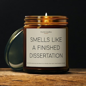 Könnte beinhalten: Eine braune Glas-Kerze mit goldenem Deckel. Auf dem Etikett steht "SMELLS LIKE A FINISHED DISSERTATION". Die Kerze ist angezündet, und der Text "Feerie Candles" steht oben auf dem Etikett.