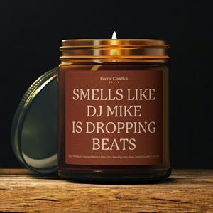 May include: A brown glass candle jar with a gold lid and a brown label featuring the text Feerie Candles, SMELLS LIKE DJ MIKE IS DROPPING BEATS, and 9oz Premium Coconut Apricot Wax, Eco-friendly, Non-toxic, Hand Poured in The USA.