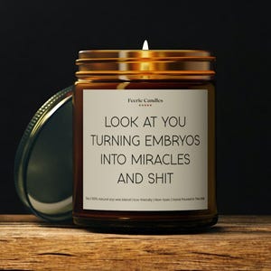 Puede incluir: Un tarro de vela de vidrio marrón con una tapa dorada. La vela tiene una etiqueta blanca con el texto "Feerie Candles" y el texto "LOOK AT YOU TURNING EMBRYOS INTO MIRACLES AND SHIT". La etiqueta también incluye el texto "100% natural soy wax blend | Eco-friendly | Non-toxic | Hand Poured in the USA".