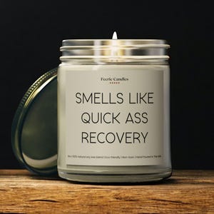 Könnte beinhalten: Eine brennende Kerze in einem durchsichtigen Glas mit einem silbernen Deckel. Auf dem Etikett steht "SMELLS LIKE QUICK ASS RECOVERY" in schwarzem Text. Das Glas steht auf einer Holzoberfläche.