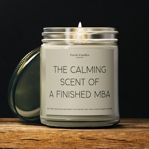 Puede incluir: Una vela en tarro de cristal con tapa plateada, etiquetada como "Feerie Candles". La etiqueta dice "THE CALMING SCENT OF A FINISHED MBA". La vela está sobre una superficie de madera. El texto en la parte inferior de la etiqueta dice "100% natural soy wax blend | Eco-friendly | Non-toxic | Hand Poured in The USA."