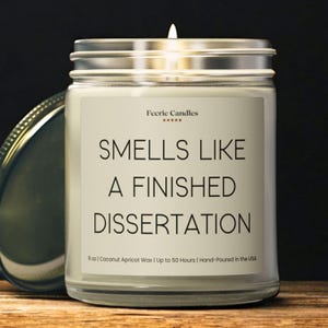 May include: A clear glass jar candle with a silver lid and a white label. The label reads Feerie Candles, five stars, and the text: SMELLS LIKE A FINISHED DISSERTATION. It includes 9 oz, Coconut Apricot Wax, Up to 50 Hours, and Hand-Poured in the USA.