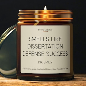 May include: A 9 oz amber glass jar candle with a gold lid and a white label. The label reads Feerie Candles, Smells Like Dissertation Defense Success, Dr. Emily, 9 oz | Coconut Apricot Wax | Up to 50 Hours | Hand-Poured in the USA. A lit wick is visible.