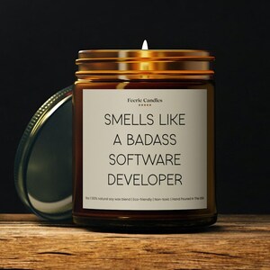 Könnte beinhalten: Eine braune Glas-Kerze mit goldenem Deckel. Auf dem Etikett steht "Smells Like A Badass Software Developer" in schwarzer Schrift. Die Kerze steht auf einer Holzoberfläche.
