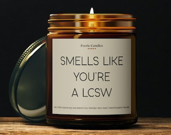 May include: A brown glass jar candle with a gold lid and a white label. The label reads "SMELLS LIKE YOU'RE A LCSW." The candle is lit, and the text "Feerie Candles" is at the top of the label.