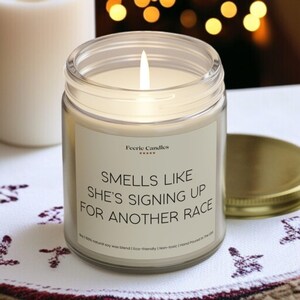 May include: A lit candle in a clear glass jar with a gold lid. The label reads "Smells Like She's Signing Up For Another Race." The candle is made with a natural soy wax blend and is hand-poured in the USA.