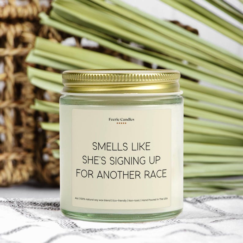 May include: A clear glass candle jar with a gold metal lid and a cream-colored label. The label features the text Feeric Candles and the phrase SMELLS LIKE SHE'S SIGNING UP FOR ANOTHER RACE, with 4oz and other small text printed below.