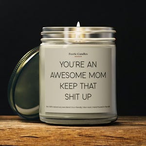 May include: A lit candle in a clear glass jar with a silver lid. The label reads "YOU'RE AN AWESOME MOM KEEP THAT SHIT UP" in black text. The candle is on a wooden surface.