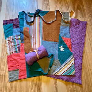 May include: A patchwork quilt made of various fabrics in shades of green, blue, brown, red, and purple. The quilt is folded and rolled up, with a small purple pillow tucked inside. The quilt is made of different patterns, including stripes, plaid, and solid colors.