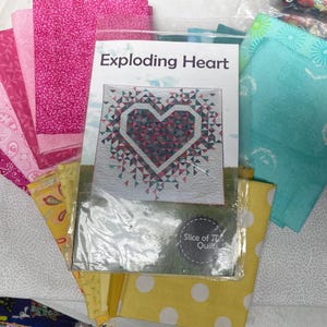 May include: A quilt pattern booklet titled Exploding Heart by Slice of Pi Quilts, featuring a heart design on the cover, surrounded by folded fabric squares in shades of pink, yellow, and teal with various patterns including paisley and polka dots.