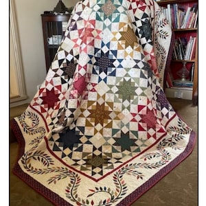 Puede incluir: Un edredón hecho a mano con un patrón de estrellas en varios colores, incluyendo rojo, azul, verde y dorado. El edredón tiene un borde color crema con un diseño de hojas y un borde rojo oscuro. El edredón está sobre una mesa.