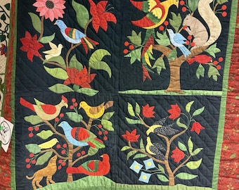 Könnte beinhalten: Eine handgefertigte Steppdecke mit dunkelblauem Hintergrund, auf der farbenfrohe Applikationen von Vögeln, Blumen und einem Eichhörnchen zu sehen sind. Die Steppdecke hat einen roten Blumenrand und grüne Akzente und zeigt einen Volkskunststil. Das Design ist in vier Quadrate unterteilt.