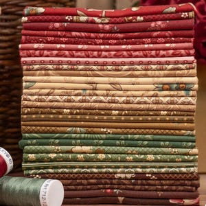 Puede incluir: Una pila de cuadrados de tela doblados en varios tonos de rojo, rosa, verde y marrón. Las telas presentan diseños florales y estampados. Una bobina de hilo verde y una bobina de hilo rojo están en primer plano.
