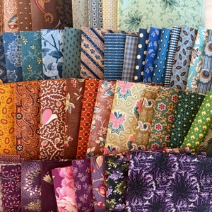 May include: A selection of fabric bundles in diverse colours and patterns. The textiles display floral, paisley, and geometric designs in shades of blue, brown, green, purple, and pink. The bundles are neatly stacked, showcasing the variety of textiles.