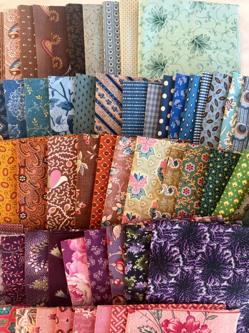 May include: A selection of fabric bundles in diverse colours and patterns. The textiles display floral, paisley, and geometric designs in shades of blue, brown, green, purple, and pink. The bundles are neatly stacked, showcasing the variety of textiles.