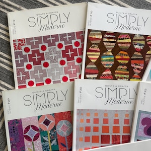 Pode incluir: Uma coleção de cinco revistas Simply Moderne Quilts & Crafts incluindo as edições 10, 19, 22, 29, 30 e 32. Cada capa apresenta um padrão de colcha único com várias cores, incluindo vermelho, laranja, castanho e estampados florais em fundo branco.
