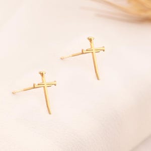 Può includere: Un paio di orecchini a forma di croce dorati. Le croci sono semplici e delicate, con una parte inferiore leggermente appuntita.