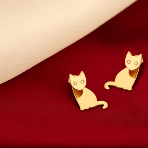May include: A pair of gold cat-shaped earrings on a red background.