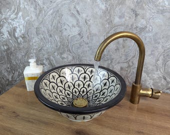 May include: A patterned ceramic sink with a black border and a white and black floral design. A brass tap is positioned above the sink, with water flowing into the basin. A soap dispenser is to the left.