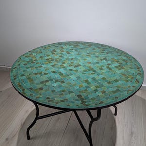May include: A round mosaic tile table with a teal and green pattern. The table has a black metal frame and legs. The table's diameter is approximately 36 inches.