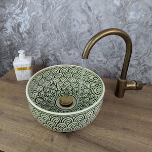 May include: A decorative ceramic sink bowl with a green and white pattern, paired with a brass tap. The sink is round and has a detailed design. A white soap dispenser with gold detailing is also visible.
