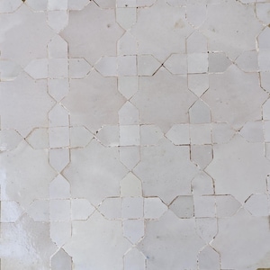Pode incluir: Close-up de um padrão de azulejos brancos. Os azulejos estão dispostos em um design geométrico repetitivo com formas de estrela. Os azulejos têm uma superfície ligeiramente texturizada e um acabamento fosco.