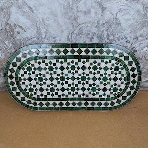 May include: An oval mosaic table featuring a green and white geometric design. The surface is composed of small, square tiles arranged in a diamond and rectangular pattern. The border is a darker green. The table is set against a neutral background.