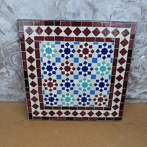 Puede incluir: Mesa cuadrada de azulejos de mosaico con un patrón geométrico. El borde presenta diamantes granates y cuadrados blancos alternados. El diseño central incluye formas de estrella y cuadrado azules, turquesas y granates. La mesa probablemente se utiliza con fines decorativos.