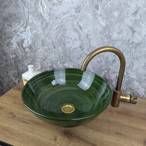 Puede incluir: Un lavabo de vidrio verde con un grifo de latón. El lavabo tiene forma circular con un patrón arremolinado verde oscuro. El grifo tiene un caño curvo. El lavabo está sobre una superficie de madera.