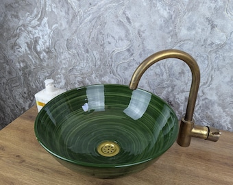 May include: A green glass vessel sink with a brass tap. The sink has a circular shape with a dark green, swirled pattern. The tap has a curved spout. The sink is on a wooden surface.