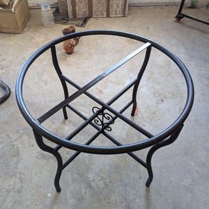 May include: A black metal outdoor table frame with a circular top and decorative scrollwork. The frame has four curved legs and a cross-shaped support structure. The table frame is designed to hold a tabletop, suitable for a patio or garden.