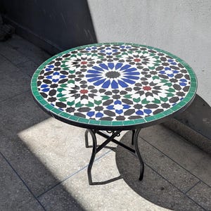Peut inclure: Table ronde en mosaïque avec une base en métal noir. Le plateau présente un motif géométrique coloré en bleu, vert, noir, rouge et blanc. La table mesure environ 61 cm de diamètre.