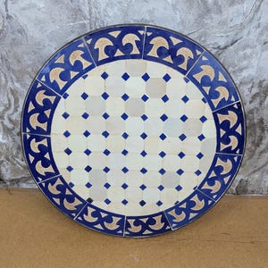 May include: Round mosaic table with a blue and white tile design. The table features a border of blue and tan decorative elements. The center of the table has a grid pattern of white and blue tiles. The table is approximately 30 inches in diameter.