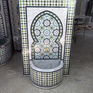 May include: A mosaic tile fountain with an arched design and a basin. The fountain has a gold tap and is decorated with green, white, and blue tiles. Other mosaic tile panels are visible in the background, showcasing a variety of patterns.