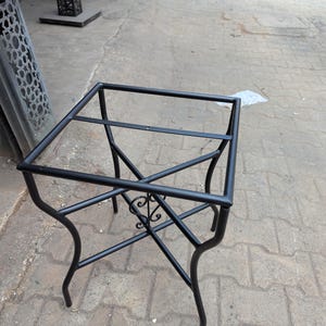 May include: A black metal table base with a square top and curved legs. The base features decorative scrollwork in the center and is designed to support a tabletop. The frame is made of black metal.