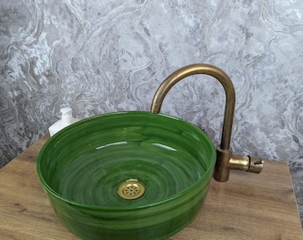 May include: A round, green ceramic bathroom sink with a brass faucet. The sink has a glossy finish and a decorative drain. The faucet is curved and has a vintage look. The sink is placed on a wooden surface.