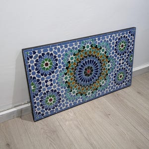 May include: A rectangular mosaic tile artwork with a geometric pattern in blue, green, and brown. The design features a central circular motif surrounded by floral shapes. The tile is framed in black and is against a white wall.